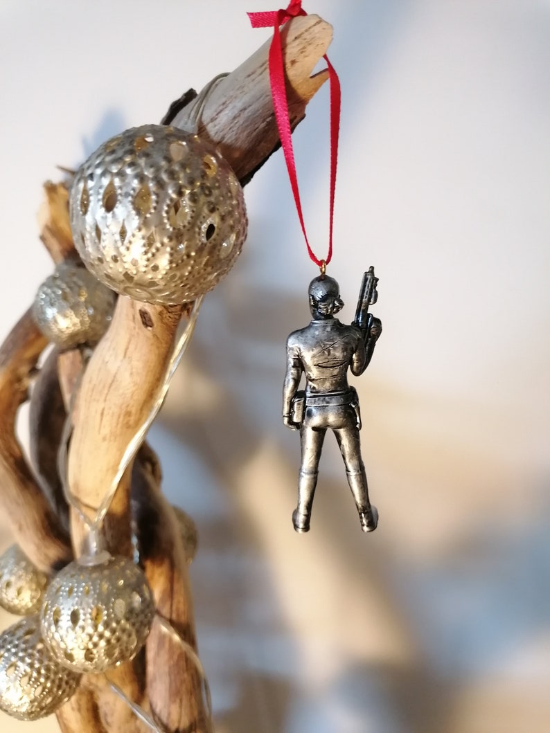 May include: A silver-coloured ornament of a person holding a gun, hanging from a red ribbon on a branch with gold-coloured ornaments.