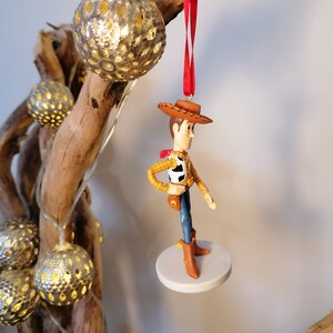 Op de afbeelding: Een rood touwtje hangt aan een tak met een witte ronde basis en een beeldje van Woody uit Toy Story dat erop staat. Woody draagt zijn kenmerkende bruine hoed, gele vest en blauwe spijkerbroek.