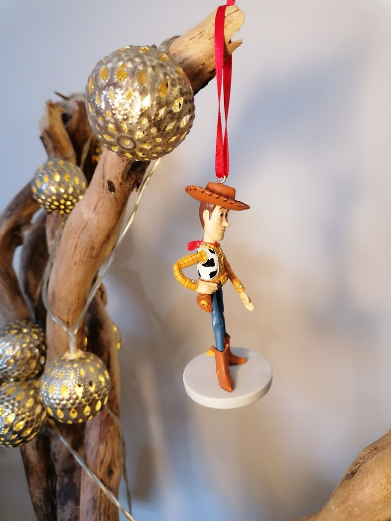 Op de afbeelding: Een rood touwtje hangt aan een tak met een witte ronde basis en een beeldje van Woody uit Toy Story dat erop staat. Woody draagt zijn kenmerkende bruine hoed, gele vest en blauwe spijkerbroek.