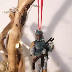 May include: A green and brown Star Wars Boba Fett Christmas ornament with a red ribbon hanging from a wooden branch.