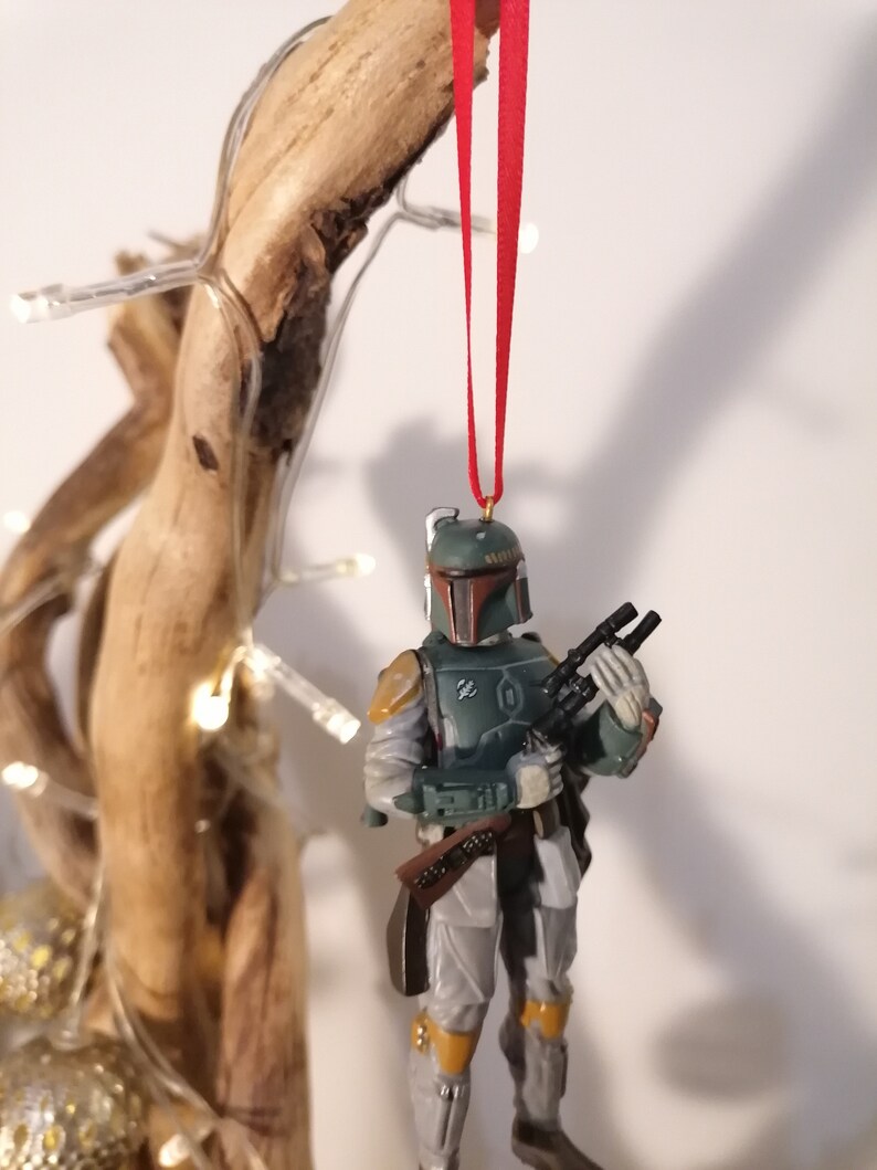 May include: A green and brown Star Wars Boba Fett Christmas ornament with a red ribbon hanging from a wooden branch.