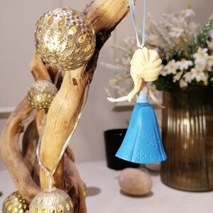 May include: A blue and white plastic ornament of a Disney princess with blonde hair and a blue dress. The ornament is hanging from a blue ribbon on a wooden branch.