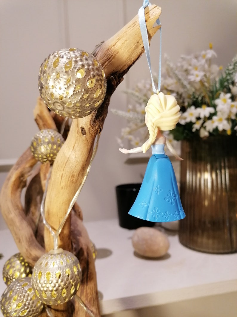 May include: A blue and white plastic ornament of a Disney princess with blonde hair and a blue dress. The ornament is hanging from a blue ribbon on a wooden branch.