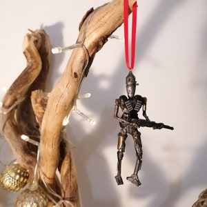 Puede incluir: Un adorno de robot gris met&aacute;lico con una cinta roja colgando de una rama. El robot sostiene un arma y tiene un dise&ntilde;o detallado.