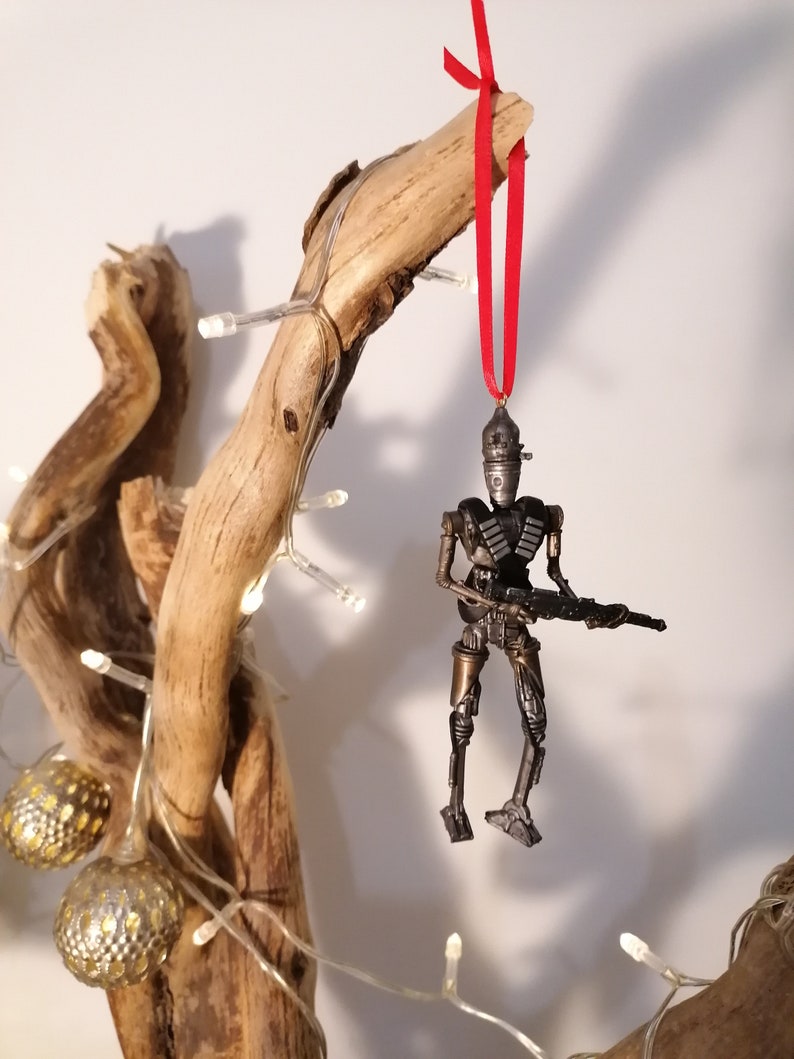 Puede incluir: Un adorno de robot gris met&aacute;lico con una cinta roja colgando de una rama. El robot sostiene un arma y tiene un dise&ntilde;o detallado.