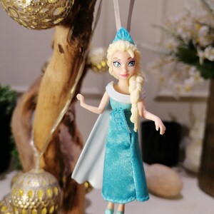 May include: A blue and white plastic doll with blonde hair wearing a blue dress and a white cape. The doll is hanging from a white string.