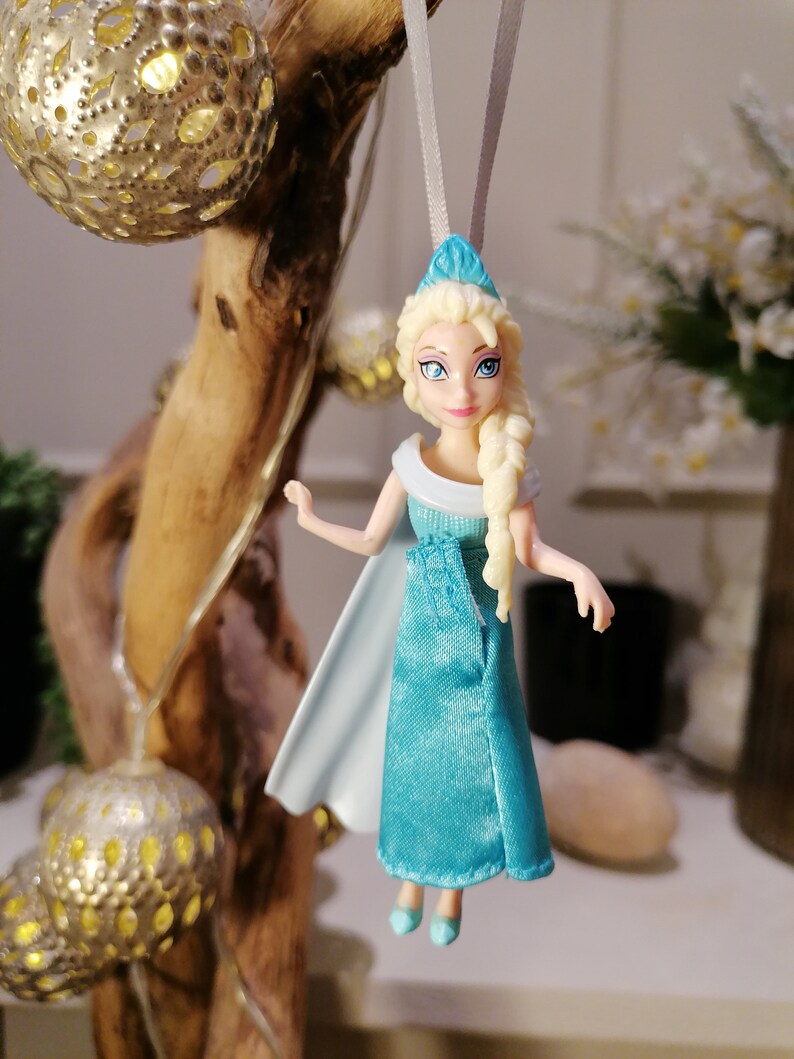 May include: A blue and white plastic doll with blonde hair wearing a blue dress and a white cape. The doll is hanging from a white string.