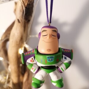 K&ouml;nnte beinhalten: Ein gr&uuml;ner und violetter Plastik-Anh&auml;nger von Buzz Lightyear aus der Toy Story-Reihe. Er tr&auml;gt seine Space-Ranger-Uniform und hat die Augen geschlossen. Der Anh&auml;nger h&auml;ngt an einer violetten Schnur.