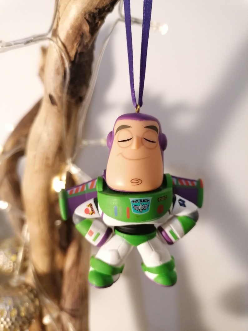 K&ouml;nnte beinhalten: Ein gr&uuml;ner und violetter Plastik-Anh&auml;nger von Buzz Lightyear aus der Toy Story-Reihe. Er tr&auml;gt seine Space-Ranger-Uniform und hat die Augen geschlossen. Der Anh&auml;nger h&auml;ngt an einer violetten Schnur.
