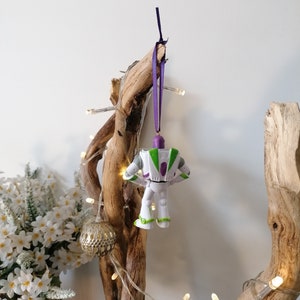May include: A white plastic toy figurine of Buzz Lightyear from the Toy Story movie franchise hanging from a purple ribbon on a branch of driftwood with white fairy lights.