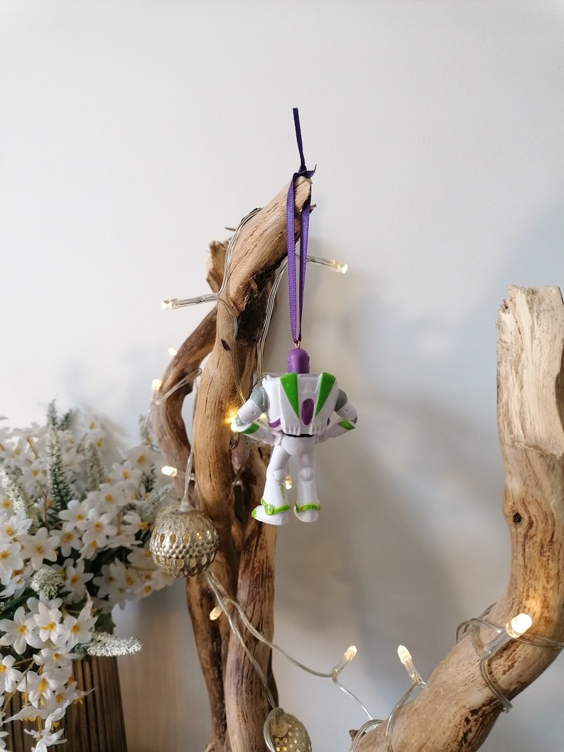 May include: A white plastic toy figurine of Buzz Lightyear from the Toy Story movie franchise hanging from a purple ribbon on a branch of driftwood with white fairy lights.