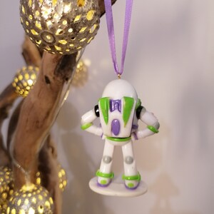 Puede incluir: Un adorno navide&ntilde;o de Buzz Lightyear blanco y verde con una pieza de pecho morada y una base morada y verde. El adorno est&aacute; colgado de una cinta morada.