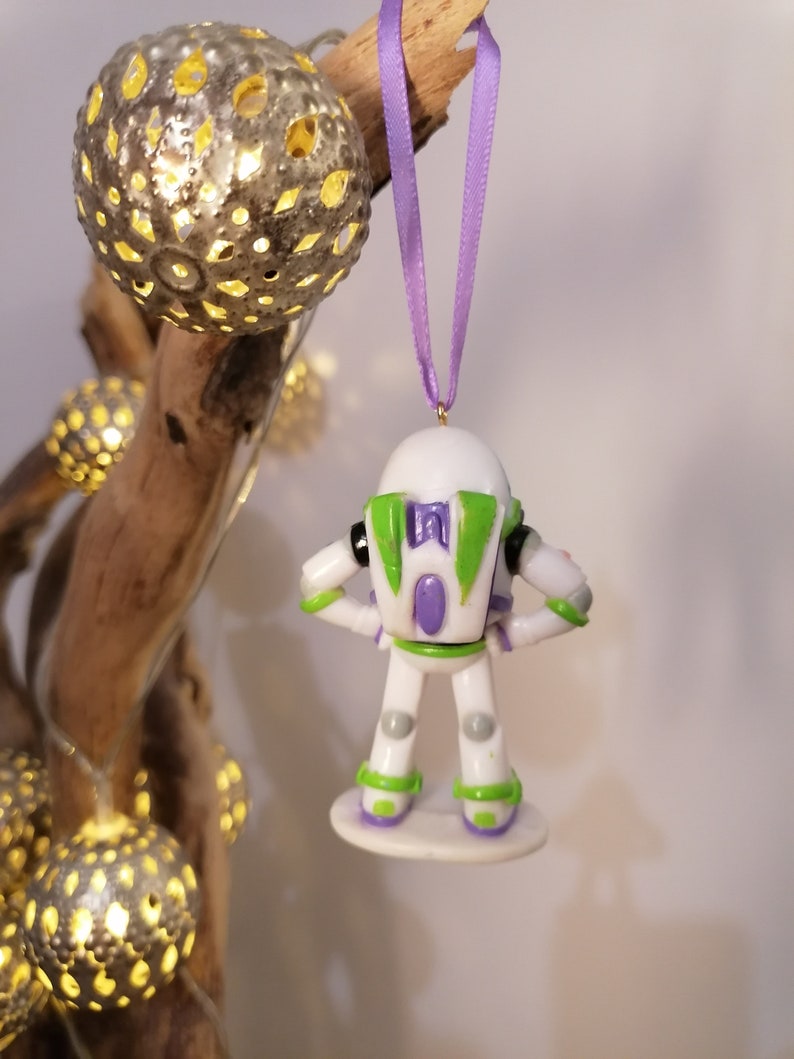 Puede incluir: Un adorno navide&ntilde;o de Buzz Lightyear blanco y verde con una pieza de pecho morada y una base morada y verde. El adorno est&aacute; colgado de una cinta morada.