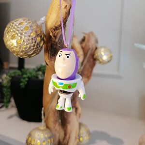 Puede incluir: Adorno navide&ntilde;o morado y blanco con un personaje de dibujos animados con cabeza morada y cuerpo blanco. El personaje lleva un traje de ranger espacial con un chaleco verde y un c&iacute;rculo rojo en el pecho. El adorno est&aacute; colgado de una cinta morada.