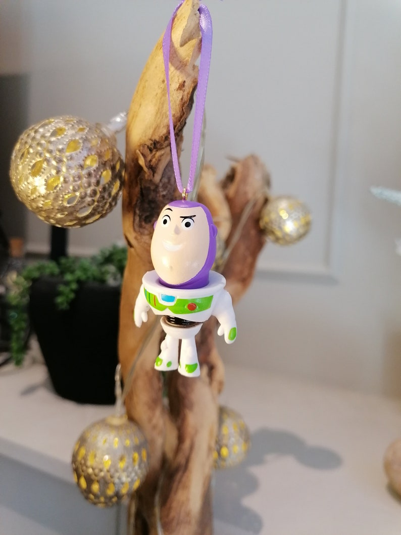 Puede incluir: Adorno navide&ntilde;o morado y blanco con un personaje de dibujos animados con cabeza morada y cuerpo blanco. El personaje lleva un traje de ranger espacial con un chaleco verde y un c&iacute;rculo rojo en el pecho. El adorno est&aacute; colgado de una cinta morada.