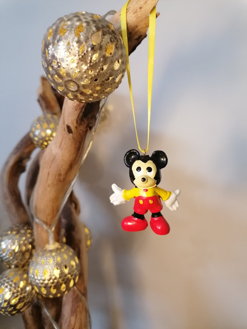 K&ouml;nnte beinhalten: Eine gelbe Schnur h&auml;ngt an einem Ast mit einem kleinen Mickey Mouse-Anh&auml;nger aus Plastik. Der Anh&auml;nger ist schwarz, gelb und rot.