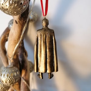 May include: Gold-colored ornament of a person wearing a cape, hanging from a red ribbon.