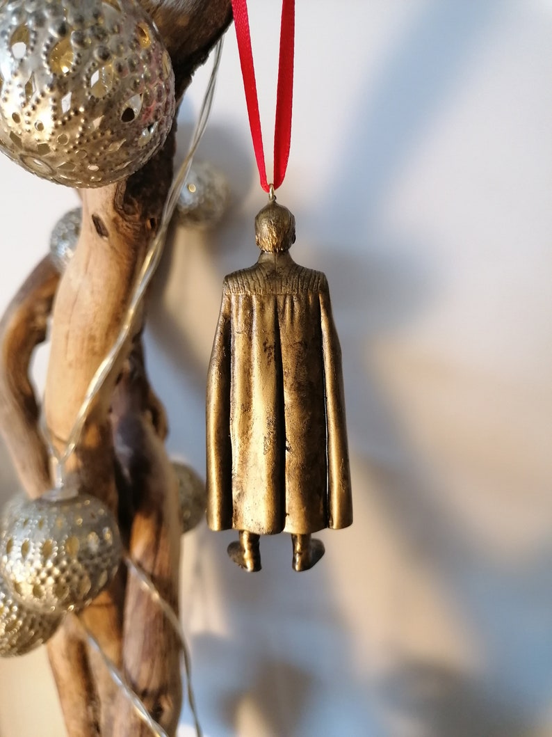 May include: Gold-colored ornament of a person wearing a cape, hanging from a red ribbon.