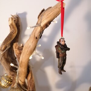 K&ouml;nnte beinhalten: Eine Miniaturfigur einer Person in milit&auml;rischer Kleidung, die an einem roten Band h&auml;ngt. Die Figur ist braun, gr&uuml;n und schwarz, mit einem Rucksack. Der Schmuck h&auml;ngt an einem Treibholzst&uuml;ck, das mit Lichterketten und goldfarbenen Kugellichtern verziert ist.