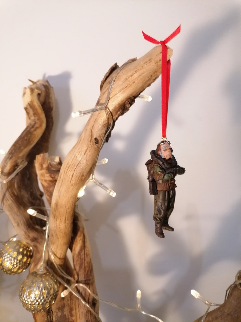 K&ouml;nnte beinhalten: Eine Miniaturfigur einer Person in milit&auml;rischer Kleidung, die an einem roten Band h&auml;ngt. Die Figur ist braun, gr&uuml;n und schwarz, mit einem Rucksack. Der Schmuck h&auml;ngt an einem Treibholzst&uuml;ck, das mit Lichterketten und goldfarbenen Kugellichtern verziert ist.