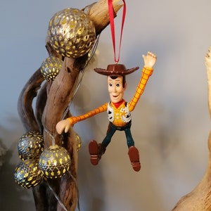 May include: A red string hangs from a wooden branch with a plastic toy figurine of Woody from Toy Story. Woody is wearing his signature cowboy hat, vest, and jeans. He is holding his arm up in the air and has a friendly expression on his face.