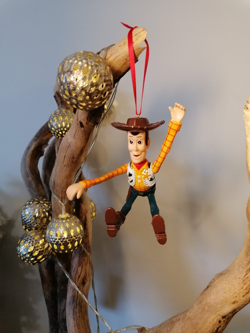 May include: A red string hangs from a wooden branch with a plastic toy figurine of Woody from Toy Story. Woody is wearing his signature cowboy hat, vest, and jeans. He is holding his arm up in the air and has a friendly expression on his face.