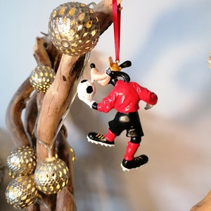 以下が含まれることがあります： 赤いサッカーボールを持った赤い服を着たGoofyのクリスマスオーナメント。Goofyは赤いジャージと黒いショートパンツを履いています。彼は右足でサッカーボールを蹴っています。