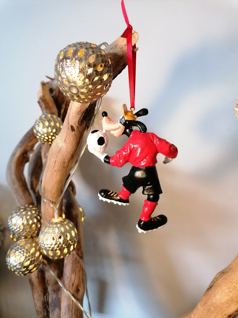 以下が含まれることがあります： 赤いサッカーボールを持った赤い服を着たGoofyのクリスマスオーナメント。Goofyは赤いジャージと黒いショートパンツを履いています。彼は右足でサッカーボールを蹴っています。
