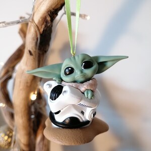 K&ouml;nnte beinhalten: Ein gr&uuml;ner und brauner Weihnachtsbaumschmuck mit einer kleinen, gr&uuml;nen Alienfigur, die in einem wei&szlig;en und schwarzen Raumschiff sitzt. Der Schmuck h&auml;ngt an einem gr&uuml;nen Band.