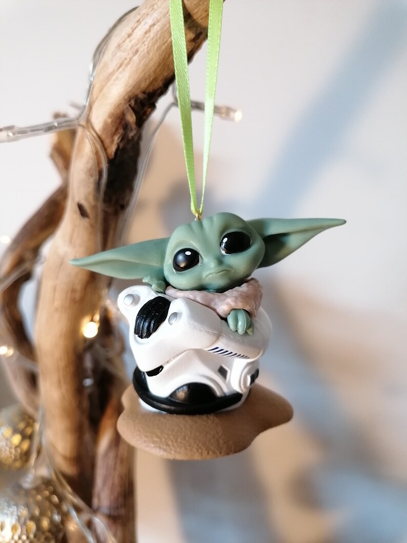 K&ouml;nnte beinhalten: Ein gr&uuml;ner und brauner Weihnachtsbaumschmuck mit einer kleinen, gr&uuml;nen Alienfigur, die in einem wei&szlig;en und schwarzen Raumschiff sitzt. Der Schmuck h&auml;ngt an einem gr&uuml;nen Band.