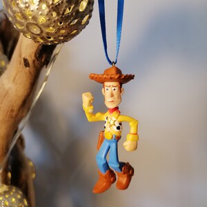 May include: A plastic toy ornament of Woody from Toy Story. Woody is wearing his signature cowboy hat, vest, and jeans. He is holding his arm up in the air, as if he is about to shoot his gun. The ornament is hanging from a blue ribbon.