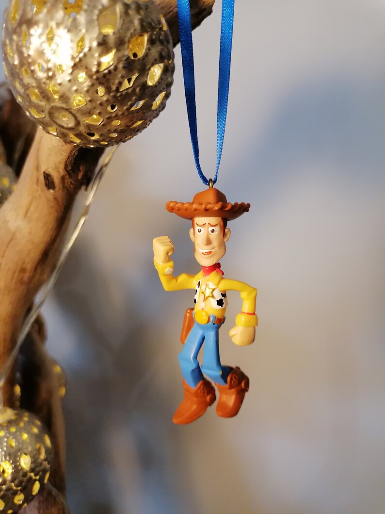 May include: A plastic toy ornament of Woody from Toy Story. Woody is wearing his signature cowboy hat, vest, and jeans. He is holding his arm up in the air, as if he is about to shoot his gun. The ornament is hanging from a blue ribbon.