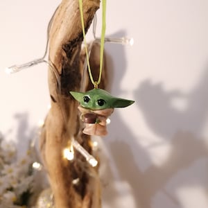 May include: A green and brown Yoda ornament with a green ribbon hanging from a wooden branch. The ornament is a popular character from the Star Wars franchise.