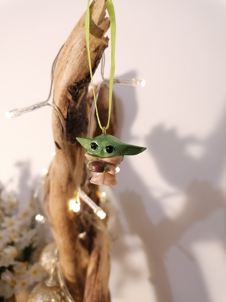 May include: A green and brown Yoda ornament with a green ribbon hanging from a wooden branch. The ornament is a popular character from the Star Wars franchise.