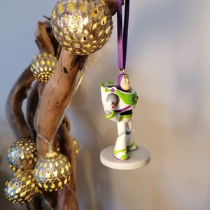 May include: A white and green Buzz Lightyear Christmas ornament with a purple ribbon hanging from a branch with silver and gold decorative lights.