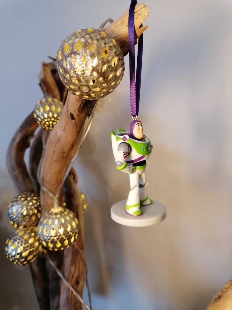May include: A white and green Buzz Lightyear Christmas ornament with a purple ribbon hanging from a branch with silver and gold decorative lights.