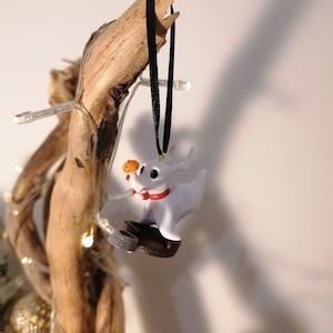 May include: White plastic ornament of Zero, the ghost dog from The Nightmare Before Christmas, holding a pumpkin. Zero is wearing a red collar and is standing on a brown base. The ornament is hanging from a black ribbon.