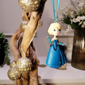 Peut inclure: Une d&eacute;coration en plastique bleu repr&eacute;sentant une princesse aux cheveux blonds et une robe bleue suspendue &agrave; un ruban bleu sur une branche avec des ornements dor&eacute;s et argent&eacute;s.