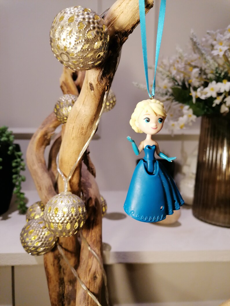 Peut inclure: Une d&eacute;coration en plastique bleu repr&eacute;sentant une princesse aux cheveux blonds et une robe bleue suspendue &agrave; un ruban bleu sur une branche avec des ornements dor&eacute;s et argent&eacute;s.