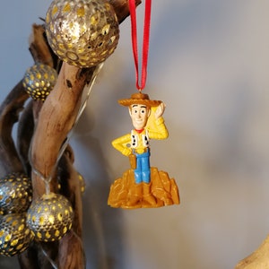 May include: A plastic ornament of Woody from Toy Story, wearing his signature cowboy hat and vest, standing on a brown base. The ornament is hanging from a red ribbon.
