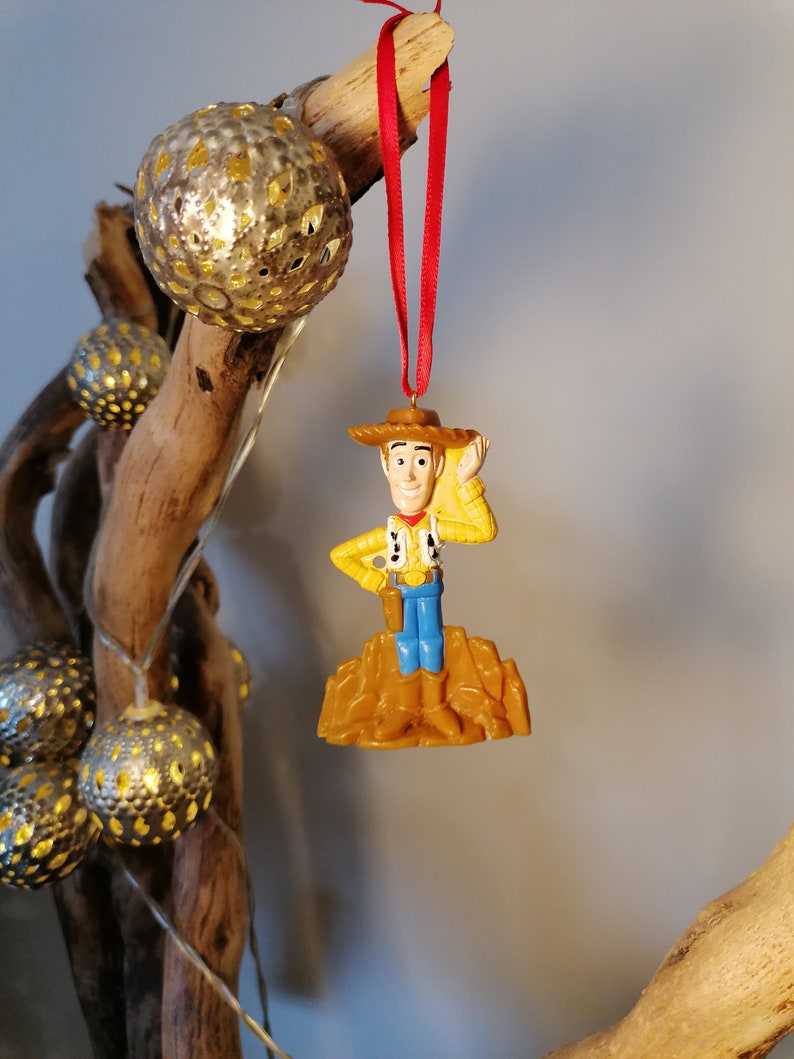 May include: A plastic ornament of Woody from Toy Story, wearing his signature cowboy hat and vest, standing on a brown base. The ornament is hanging from a red ribbon.