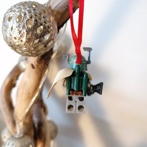 May include: A green and brown Lego Star Wars Boba Fett ornament with a red ribbon. The ornament is hanging from a piece of driftwood.