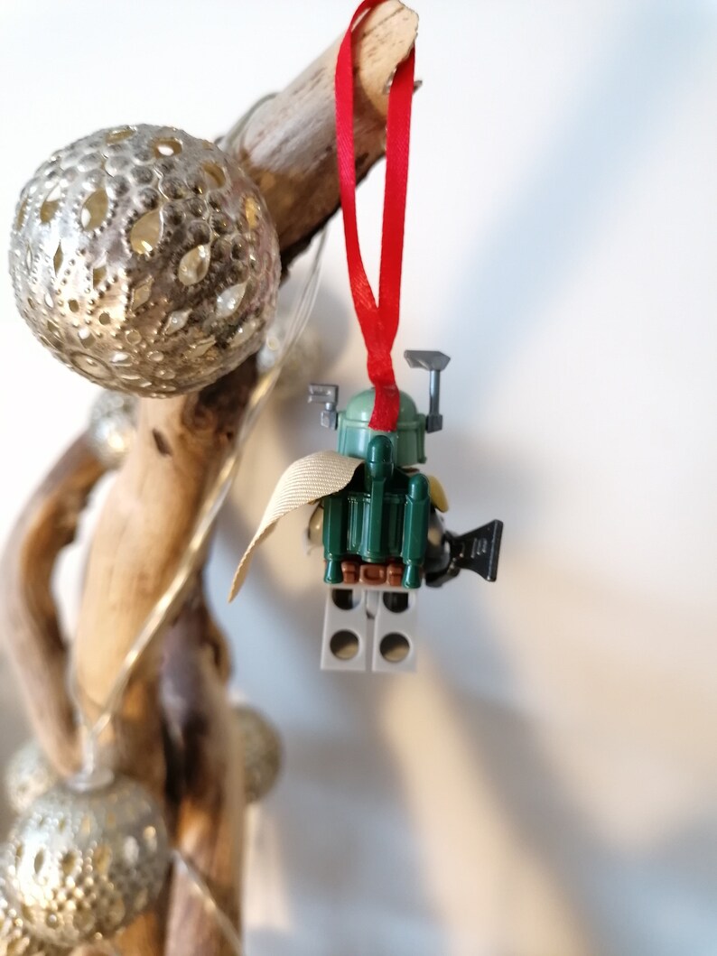 May include: A green and brown Lego Star Wars Boba Fett ornament with a red ribbon. The ornament is hanging from a piece of driftwood.