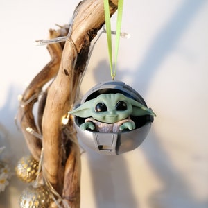 May include: A green and gray Christmas ornament featuring a character from the Star Wars franchise, The Mandalorian. The character is sitting in a pod-like structure.