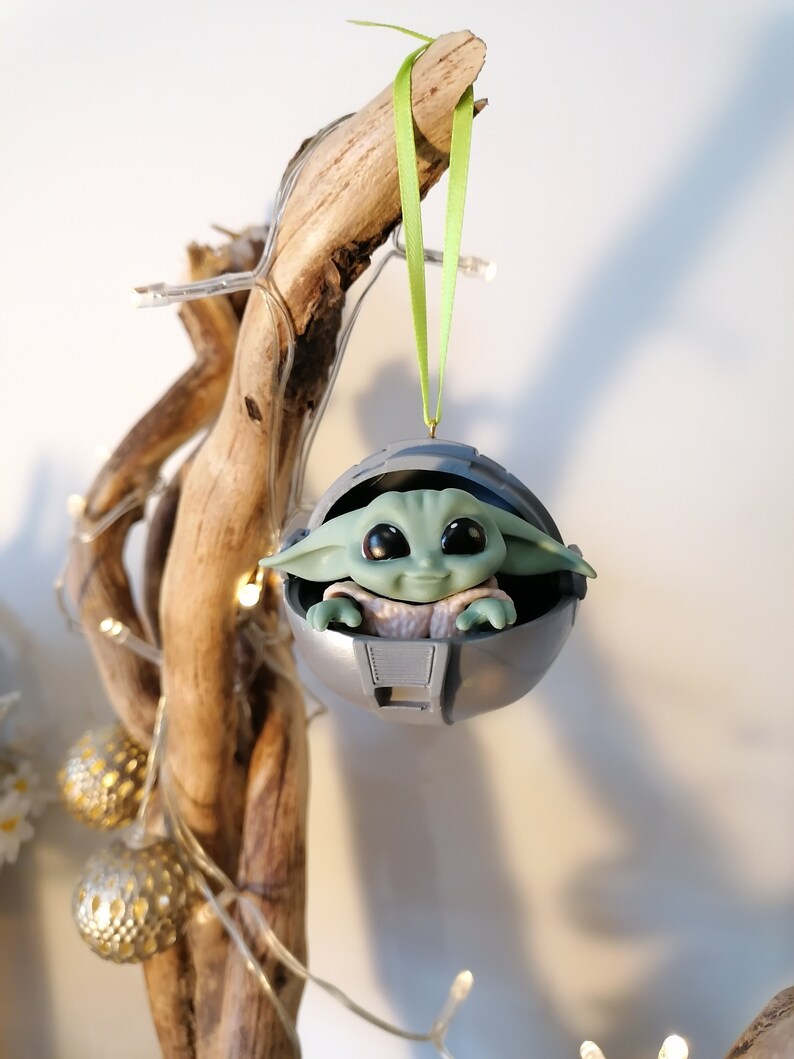 May include: A green and gray Christmas ornament featuring a character from the Star Wars franchise, The Mandalorian. The character is sitting in a pod-like structure.