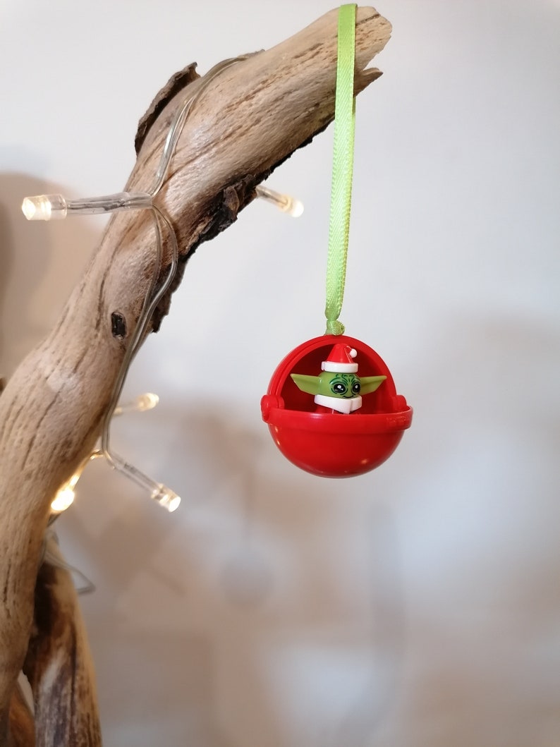 Pu&ograve; includere: Un ornamento natalizio rosso con un personaggio verde al suo interno. L'ornamento &egrave; appeso a un nastro verde su un ramo.