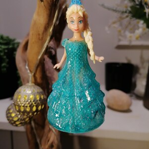 May include: A blue and white plastic doll ornament of a princess wearing a sparkly blue dress. The doll has blonde hair and blue eyes. The ornament is hanging from a string.