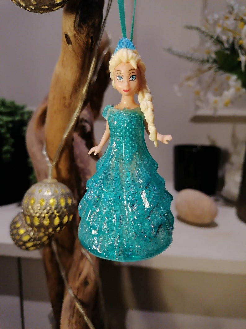 May include: A blue and white plastic doll ornament of a princess wearing a sparkly blue dress. The doll has blonde hair and blue eyes. The ornament is hanging from a string.