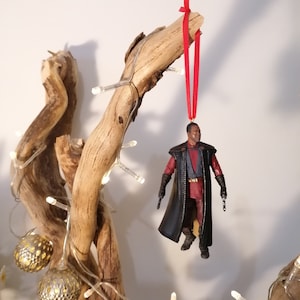 K&ouml;nnte beinhalten: Ein schwarzer und roter Star Wars-Charakter-Anh&auml;nger, der an einem Ast mit einem roten Band h&auml;ngt. Der Anh&auml;nger ist eine Figur eines Charakters, der einen schwarzen Umhang tr&auml;gt und in jeder Hand einen Blaster h&auml;lt.