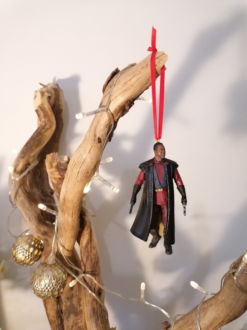 K&ouml;nnte beinhalten: Ein schwarzer und roter Star Wars-Charakter-Anh&auml;nger, der an einem Ast mit einem roten Band h&auml;ngt. Der Anh&auml;nger ist eine Figur eines Charakters, der einen schwarzen Umhang tr&auml;gt und in jeder Hand einen Blaster h&auml;lt.